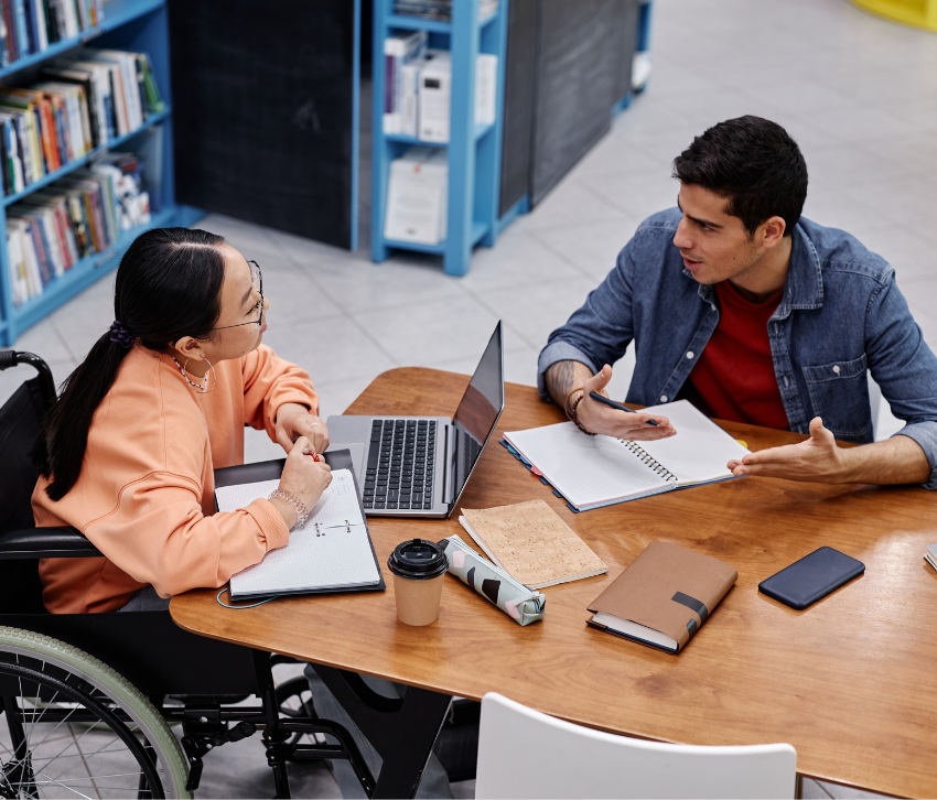 A college student in a wheelchair studies at a table in the library with another student.